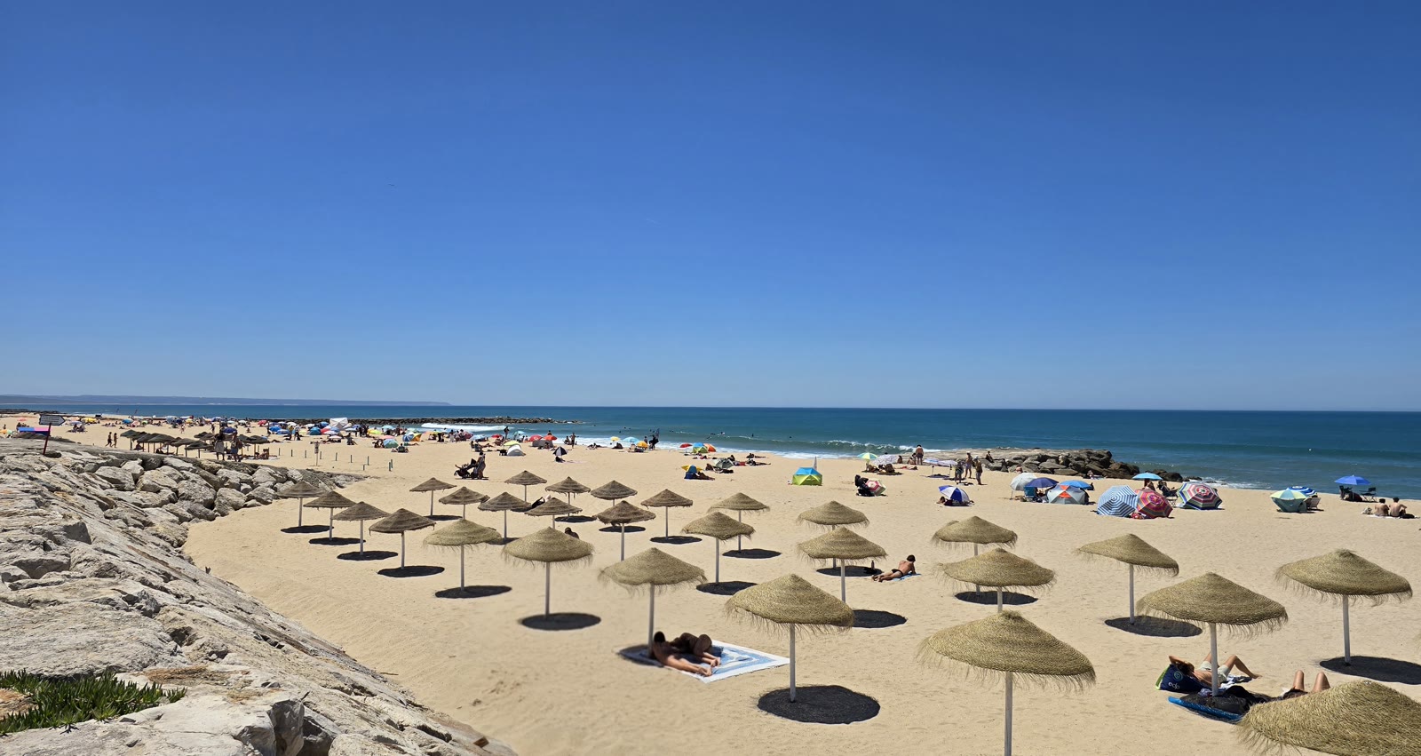 Chapéus de praia e espreguiçadeiras alinhados na areia da Costa da Caparica, mar ao fundo